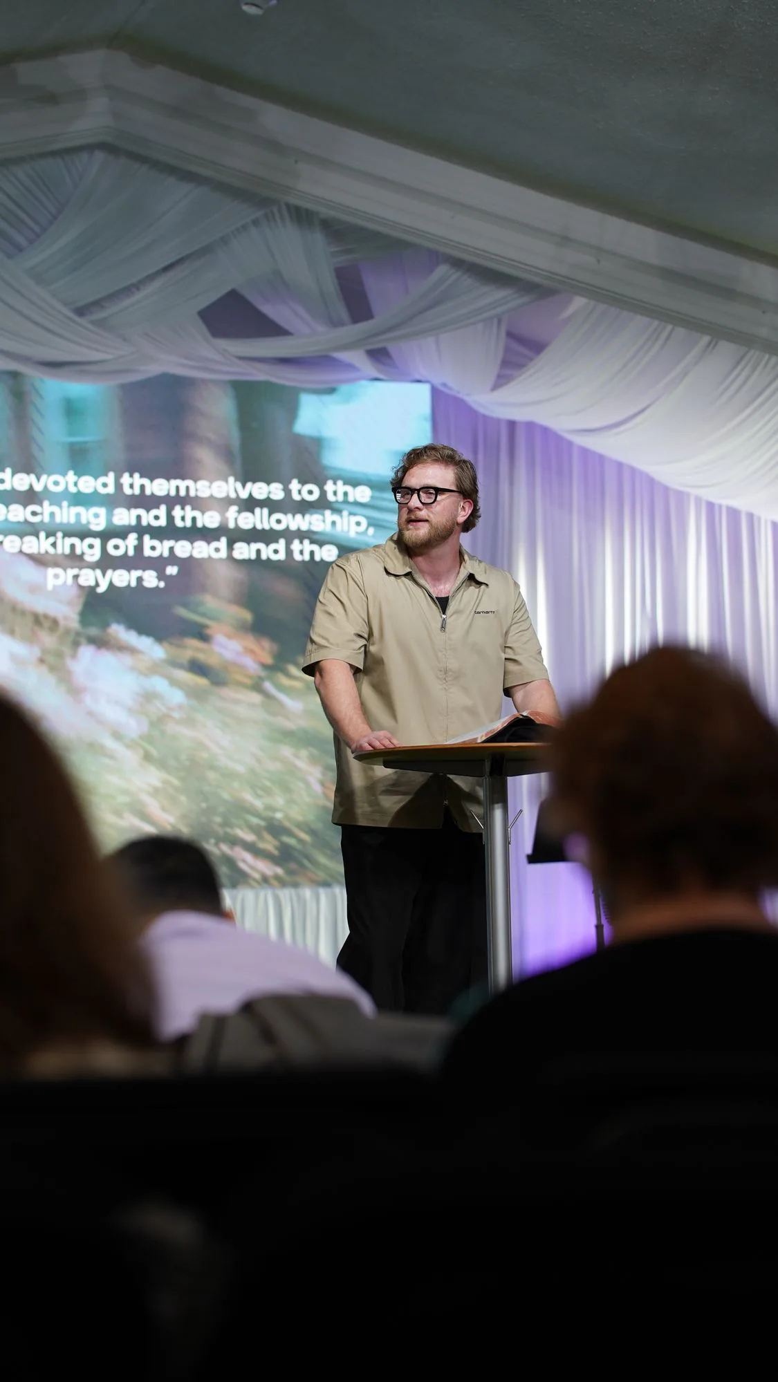 Pastor Jeremy preaching at the lectern, Acts 2:42 on the screen behind him