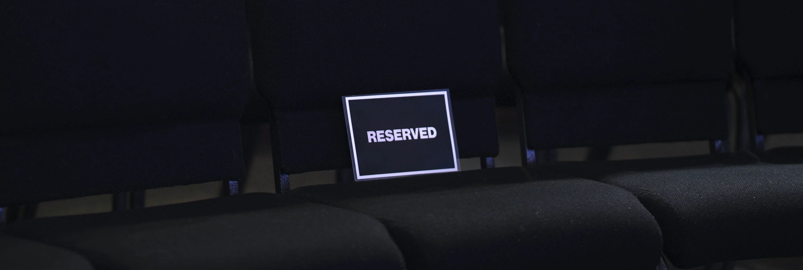 A row of empty chairs in the dark, one holding a small card that reads Reserved